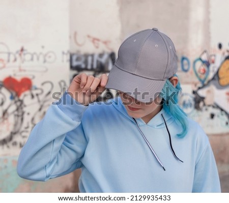 Blue haired Teenage girl in blue hoodie and baseball cap touching her cap. Blue haired teen girl stays outdoors against graffiti wall. Clothing and cap mockup 