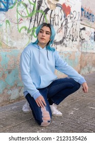 Blue Haired Teenage Girl In Light Blue Oversize Hoodie And Looking Aside. Blue Haired Teen Girl Sitting On Her Haunches Near Graffiti Wall. Clothing Mockup 