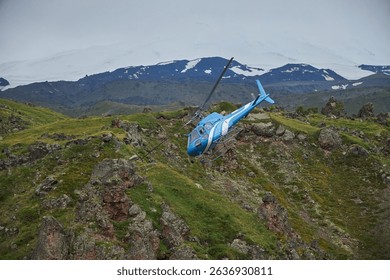 blue helicopter navigates through the dramatic landscape of Iceland, surrounded by rocky hills and a distant glacial backdrop under an overcast sky. This aerial view showcases natures beauty.
