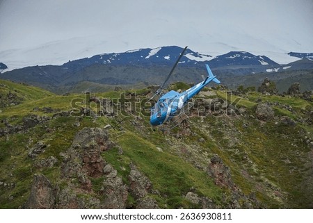 blue helicopter navigates through the dramatic landscape of Iceland, surrounded by rocky hills and a distant glacial backdrop under an overcast sky. This aerial view showcases natures beauty.
