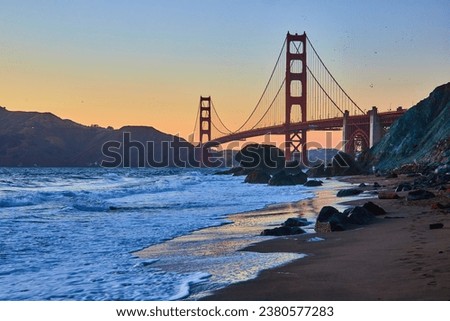 Blue hour with glowing waves against sandy shore with distant Golden Gate Bridge