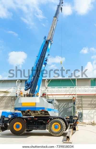blue hydraulic truck crane\
standing on a construction site under construction. A worker is\
standing on a scissor lift at a suitable height to help positioning\
it.