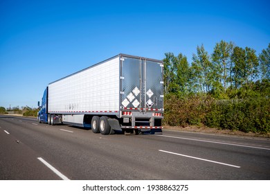 Blue industrial long haul big rig semi truck with high cabin transporting frozen cargo in refrigerator semi trailer driving on the wide multiline interstate highway road with trees on the side