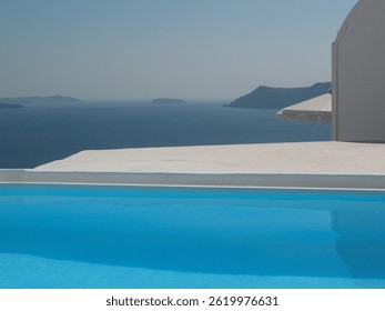 Blue infinity pool overlooking the deep blue Aegean Sea in Santorini, Greece. Bright sunlight and a minimalist Mediterranean setting create a perfect summer holiday and relaxation scene.