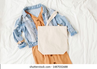 Blue Jean Jacket And  Beige Dress With Tote Bag On White Bed. Women's Stylish Autumn Outfit. Trendy Clothes With White Eco Bag Mockup. Flat Lay, Top View.