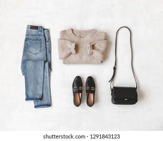 Blue Jeans, Beige Knitted Oversize Sweater, Small Cross Body Bag, Black Loafers Or Flat Shoes On Grey Background. Overhead View Of Women's Casual Day Outfit. Flat Lay, Top View. Women Clothes.