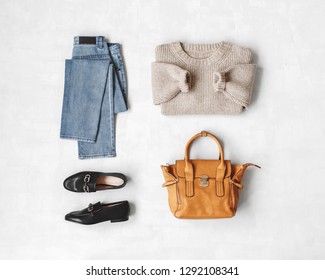 Blue Jeans, Beige Knitted Sweater, Bag, Black Loafers Or Flat Shoes On Grey Background. Overhead View Of Women's Casual Day Outfit. Flat Lay, Top View. Women Clothes.