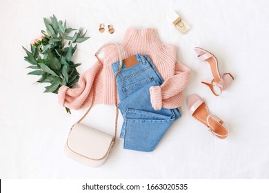 Blue jeans, pink knitted sweater, heeled sandals, small bag and bouquet of flowers lie on white background. Overhead view of woman's casual outfit. Trendy stylish women clothes. Flat lay, top view.