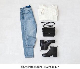 Blue jeans, white knitted sweater, small black cross body bag and leather ankle boots on grey background. Overhead view of woman's casual day outfits. Trendy hipster look. Flat lay.