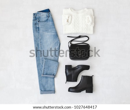 Blue jeans, white knitted sweater, small black cross body bag and leather ankle boots on grey background. Overhead view of woman's casual day outfits. Trendy hipster look. Flat lay.