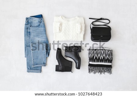 Blue jeans, white knitted sweater, small black cross body bag, leather ankle boots and striped scarf on grey background. Overhead view of woman's casual day outfits. Trendy hipster look. Flat lay.