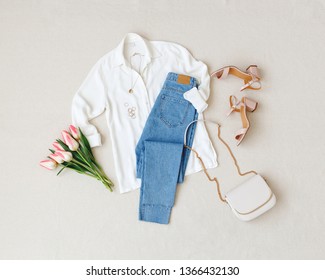 Blue jeans, white shirt, heeled sandals, bag with chain strap, jewelry, bouquet of pink tulips flowers on beige background. Women's stylish spring summer outfit. Trendy clothes. Flat lay, top view. - Shutterstock ID 1366432130