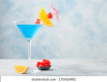 Blue Lagoon Summer Cocktail In Martini Glass With Sweet Cocktail Cherries And Orange Slice With Umbrella On Blue Table Background. Space For Text