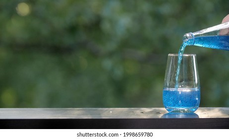 Blue liquid with pieces of jelly is poured from a bottle into a transparent glass. Making drinks outdoors. Blue drink with fruit pieces. Wooden table with green summer foliage bokeh background.