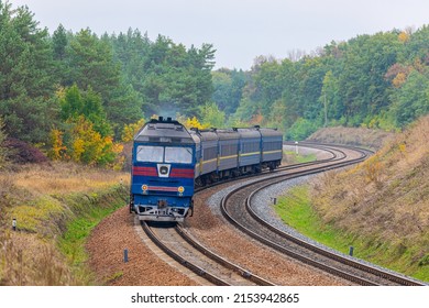 A Blue Locomotive Pulls A Passenger Train To The Railway Station Along A Winding Road. Trees With Green And Yellow Leaves. Evening Autumn Lighting.