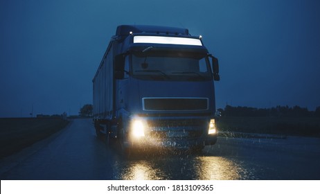 Blue Long Haul Semi-Truck with Cargo Trailer Full of Goods Travels At Night on the Freeway Road, Driving Across Continent Through Rain, Fog, Snow. Industrial Warehouses Area. Front Shot