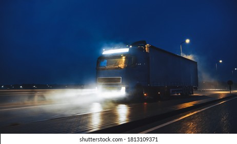 Blue Long Haul Semi-Truck with Cargo Trailer Full of Goods Travels At Night on the Freeway Road, Driving Across Continent Through Rain, Fog, Snow. Industrial Warehouses Area. Front Shot