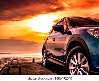 Blue luxury SUV car parked on concrete road by sea beach with beautiful red sunset sky. Summer vacation at tropical beach. Road trip. Front view sports and modern design SUV car. Summer travel by car.