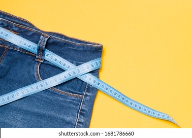 Blue Measuring Tape Is Threaded Into Jeans Instead Of A Belt, Blue Jeans On A Yellow Background, Copy Space, Weight Loss And Weight Control Concept, Measuring Body Volume