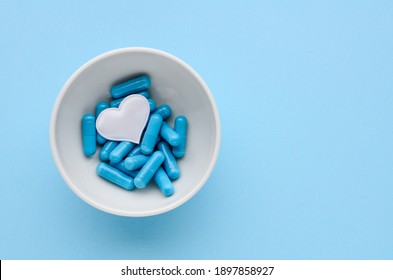 Blue medical capsules in white bowl and a white heart on a blue background. Concept of prevention and treatment of cardiovascular diseases, Valentine's day. Medical flat lay with copy space.