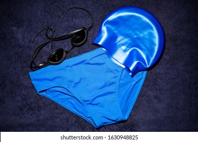 Blue men's swim gear on a navy blue towel. Swim briefs, swim cap, and swim goggles.
