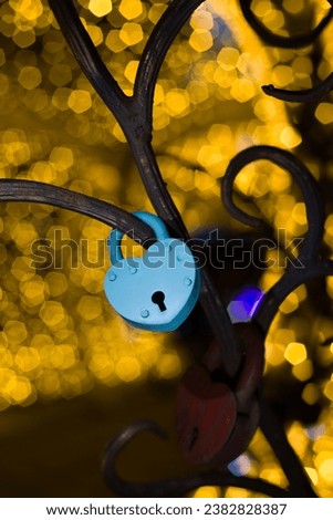 Blue metal love lock in shape of heart, as a symbol of an eternal relationship, hangs on a metal tree against a background of bright lights in the park in the evening.