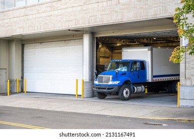 Blue Middle Class Relocation Rig Day Cab Semi Truck Tractor With Long Spacious Box Trailer Standing In Dock Inside Of The Multilevel Apartment Building In Urban City Unloading Delivered Goods