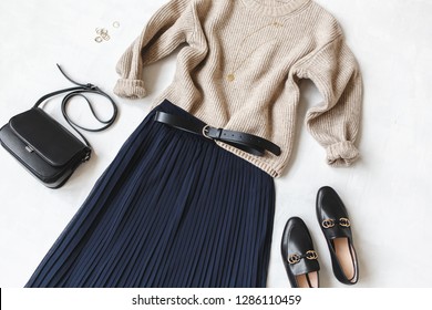 Blue Midi Pleated Skirt, Beige Knitted Sweater, Small Black Cross Body Bag, Belt, Loafers (flat Shoes) On Grey Background. Overhead View Of Women's Casual Day Outfit. Flat Lay, Top View. Women Clothes