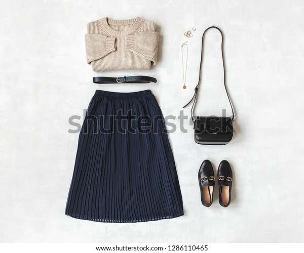 Blue midi pleated skirt, beige knitted sweater,\
small black cross body bag, belt, loafers (flat shoes) on grey\
background. Overhead view of women\'s casual day outfit. Flat lay,\
top view. Women clothes