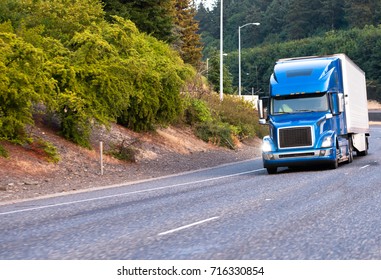 Blue Modern Popular Huge Comfortable Big Rig Semi Truck With Turn On Headlights Haul Reefer Semi Trailer For Food Delivery Moving On Wide Turning Highway With Green Trees On The Shoulder