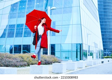 Blue Monday Concept. Young Executive Business Woman Playing With A Red Umbrella On A Cloudy Day. She Wears A Casual Urban Look, A Red Coat, Jeans And A White Blouse.