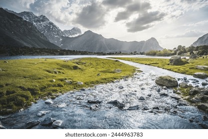 Blue mountain lake and river in a mountain valley among vegetation against the backdrop of rocky peaks with snow in the Fan Mountains in Tajikistan, Tien Shan highlands in the evening - Powered by Shutterstock