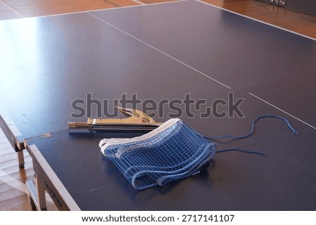 A blue net sits on a table next to a tennis racket. The net is unstrung and the racket is laying on the table. The scene suggests that someone is preparing to play tennis, but has not yet started