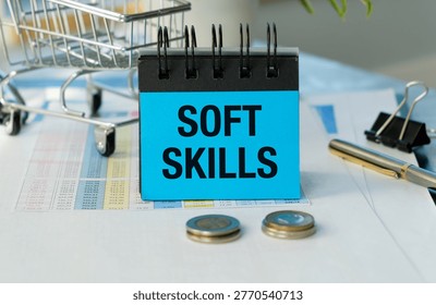 Blue notepad holds bold SOFT SKILLS text. Metal shopping cart sits nearby, symbolizing commerce. Coins rest beside the notepad, suggesting value. Paper documents and clips clutter the workspace