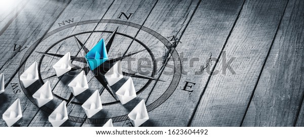 Blue\
Paper Boat Leading A Fleet Of Small White Boats With Compass Icon\
On Wooden Table With Sunlight - Leadership\
Concept