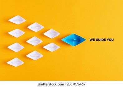 Blue paper boat leads white paper ships with the message we guide you. Guidance, support, counseling, assistance or coaching in business or education concept.