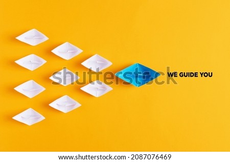 Blue paper boat leads white paper ships with the message we guide you. Guidance, support, counseling, assistance or coaching in business or education concept.