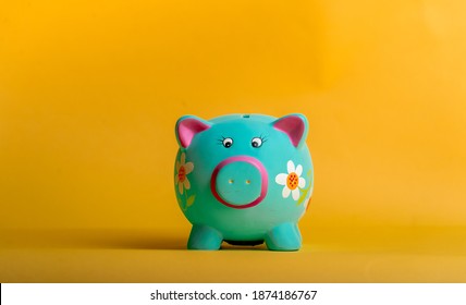 Blue Piggy Bank In Flowers Isolated On Yellow Background, Funny Bank For Kid Savings. Financial Education For Children. The Figure Of Money Savings And Investing