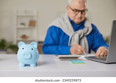 Blue piggy bank on table with a mature senior man in background calculating income and profit, finances or taxes at home, checking his bills. Pension calculation, investment and savings concept. - Powered by Shutterstock