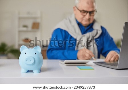Blue piggy bank on table with a mature senior man in background calculating income and profit, finances or taxes at home, checking his bills. Pension calculation, investment and savings concept.