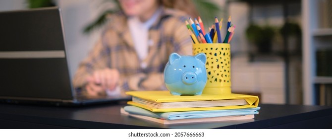 Blue Piggy Bank And Textbooks On The Table, In The Background A Teenager Girl Does Homework On A Laptop. Training Cost Concept