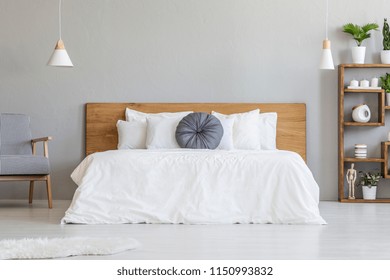 Blue pillow on white bed with wooden headboard in bedroom interior with armchair. Real photo