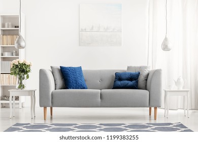 Blue Pillows On Grey Couch Next To Table With Flowers In White Flat Interior With Painting. Real Photo