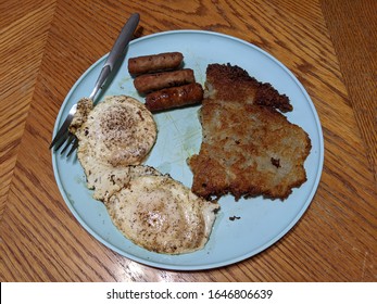 A blue plastic breakfast plate of three pork maple sausages two eggs sunny side up soft and a home made hash brown potato salted and served on a wooden table with a fork