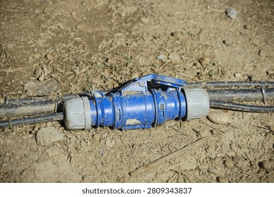A blue plastic cable connector with a locking handle joins two black cables resting on dry, cracked soil in an outdoor setting under natural light.
