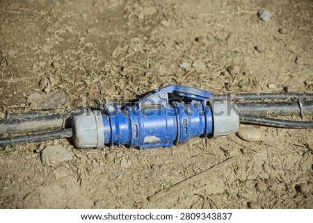 A blue plastic cable connector with a locking handle joins two black cables resting on dry, cracked soil in an outdoor setting under natural light.