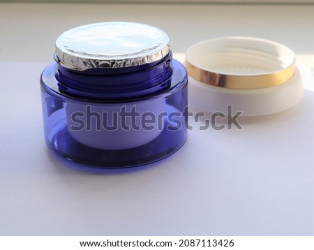 a blue plastic jar of cream with a sealed foil on top and a light lid with a gold border behind on a light background, an unused object of women's face skin care cosmetics