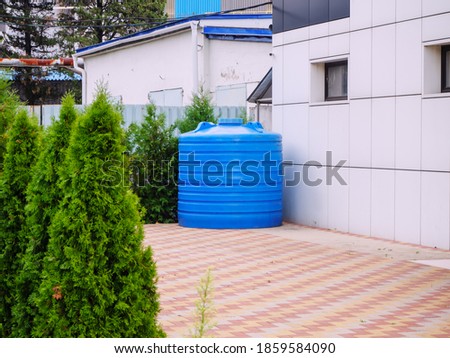 A blue plastic water tank stands in a backyard surrounded by thujas