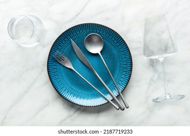 A Blue Plate With A Fork, A Knife, And A Spoon, With A Glass For Water And A Wineglass, Overhead Flat Lay Shot On A White Marble Background