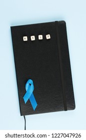 Blue Prostate Ribbon On Black Leather Notepad Isolated On Blue Background, Prostate Cancer And Symbol Of Helping HIV Patients